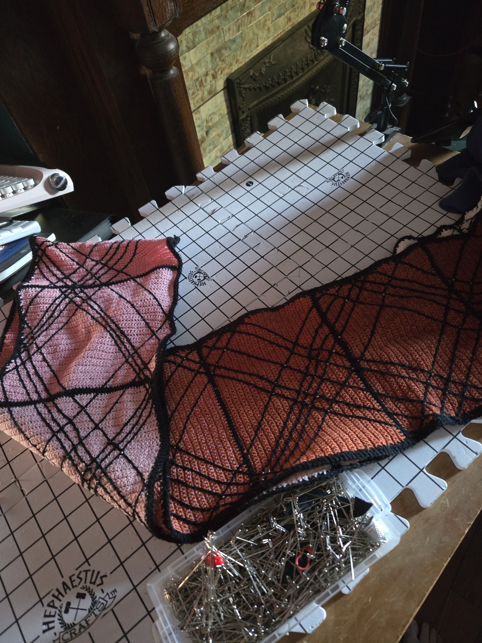 Picture of scarf on gridded blocking mats, The scarf goes from white, to pink, to rusts. Black lines across the scarf form a scale pattern.