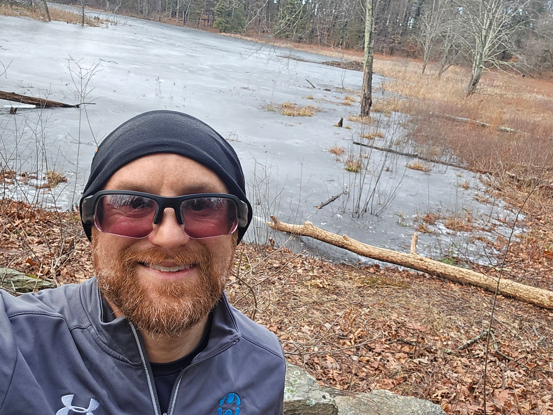 A selfie of me in front of the melting ice on top of a pond in dry, swampy land next to a forest. I'm a bald, middle-aged, white man with a red beard flecked with white. I'm wearing a black headband, black sunglasses, and a grey Under Armour running sweatshirt with the Humanyze logo on the right.