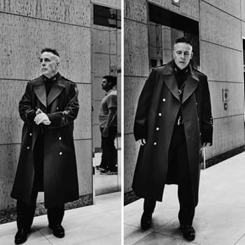 A middle-aged man with a fashy haircut and a long black overcoat stands on a city street outside a building.