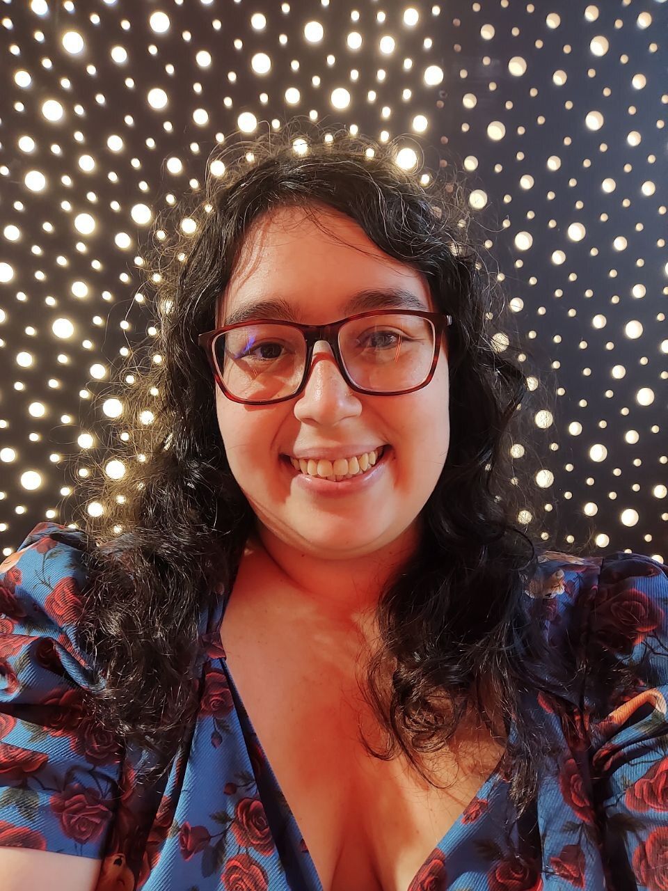 Fotografia da autora Claudia Fusco, uma mulher branca de corpo gordo e cabelos pretos cacheados. Ela usa óculos e sorri diretamente para a câmera na frente de um fundo escuro com pontinhos brilhantes.