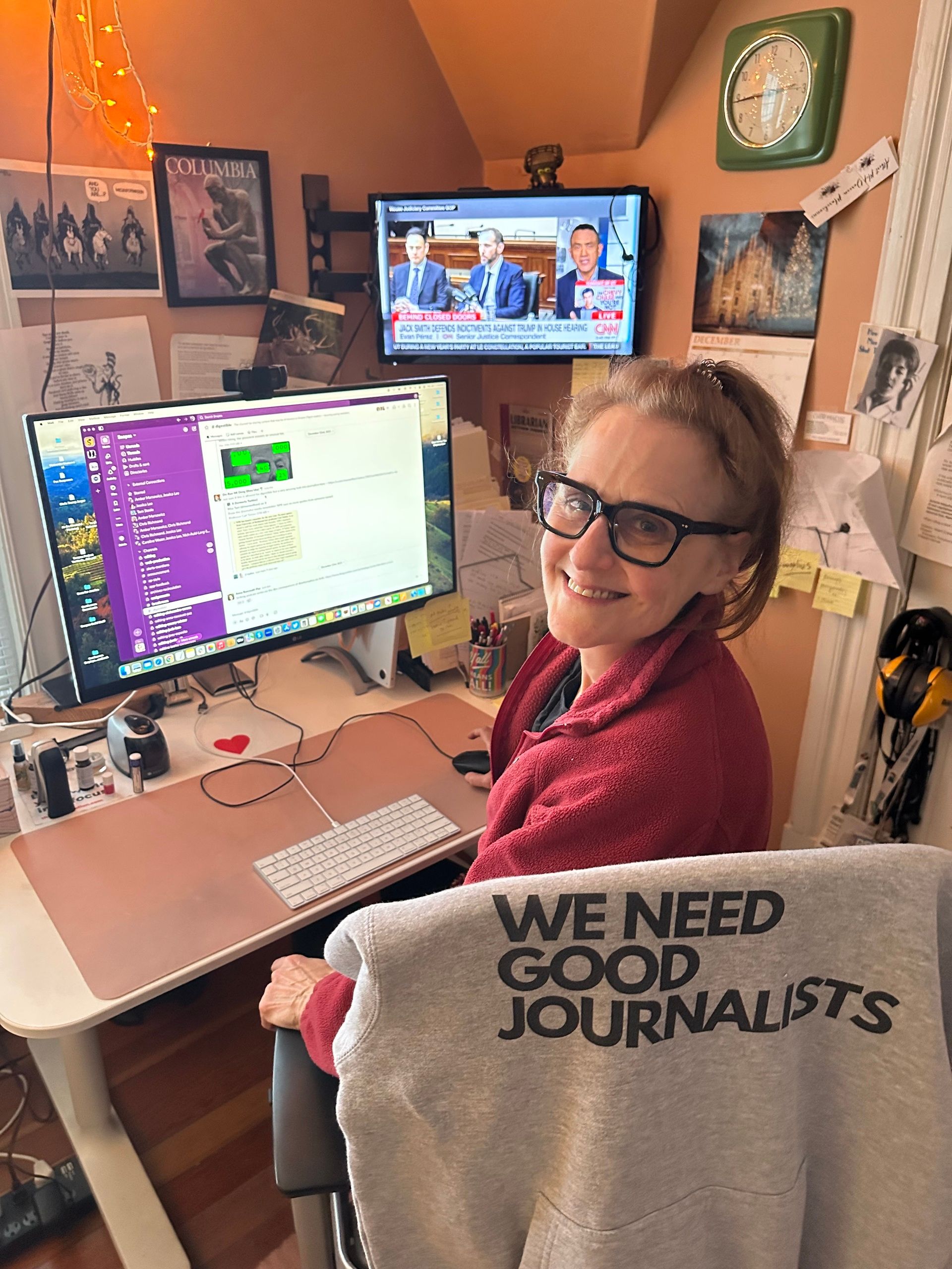 A white woman smiles while looking at the camera. Her desktop laptop shows a Slack workspace. A TV airing CNN is shown in the corner of the room, while the sweeatshirt hanging on her chair says WE NEED GOOD JOURNALISTS.