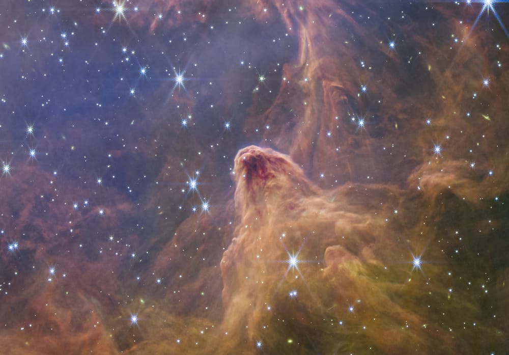 A close-up of one of the fingers of dust and gas, looking like a slightly tipped over orange-ish mountain with a black apex in a sea of orange fog and stars.