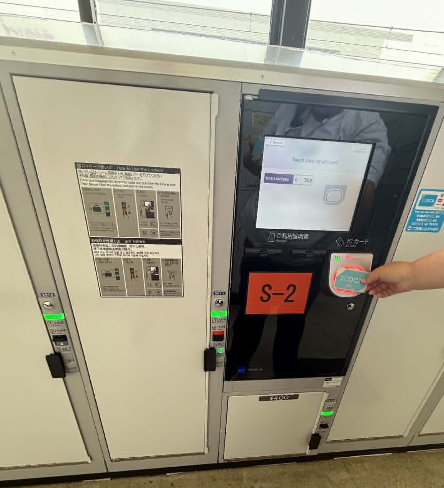 Coin-operated locker system in a Japanese train station with a digital payment screen and card reader. A hand is tapping a transit card to access the locker, showing how luggage can be stored securely while exploring.