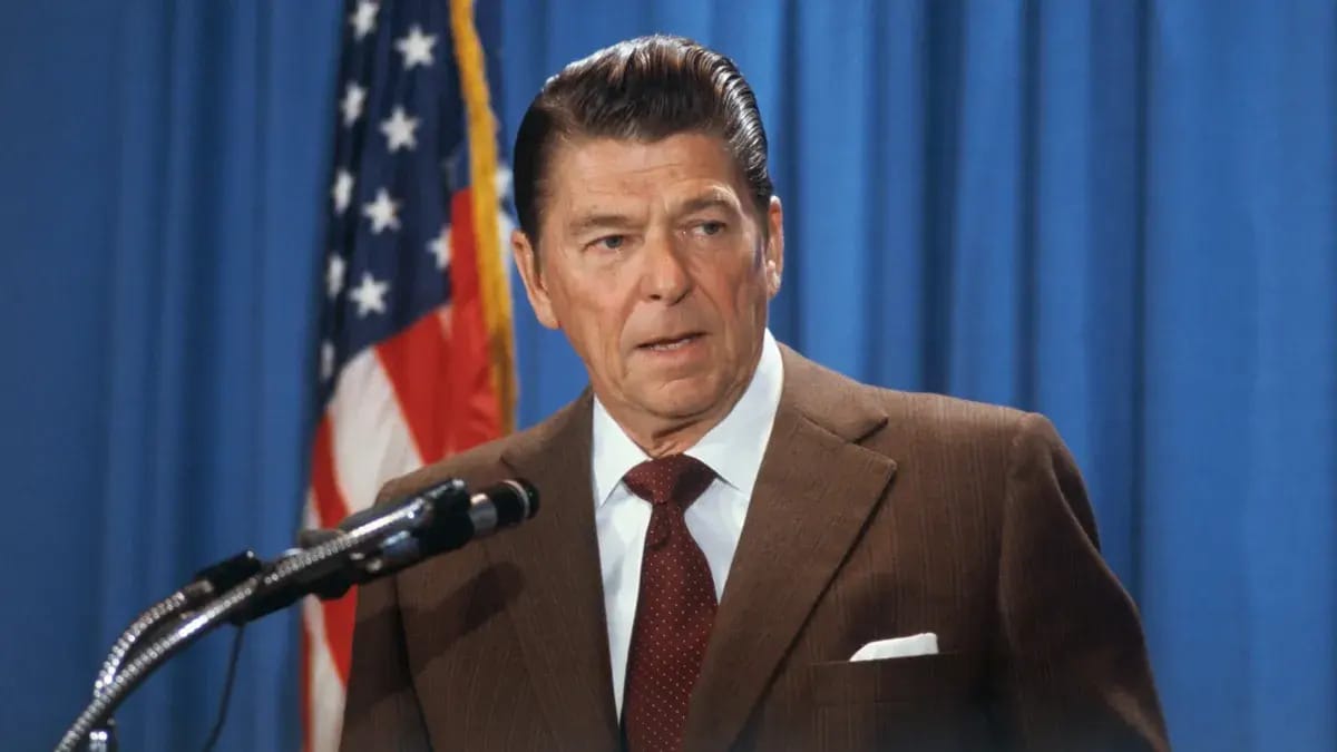 A white man wearing a brown suit stands before a microphone.