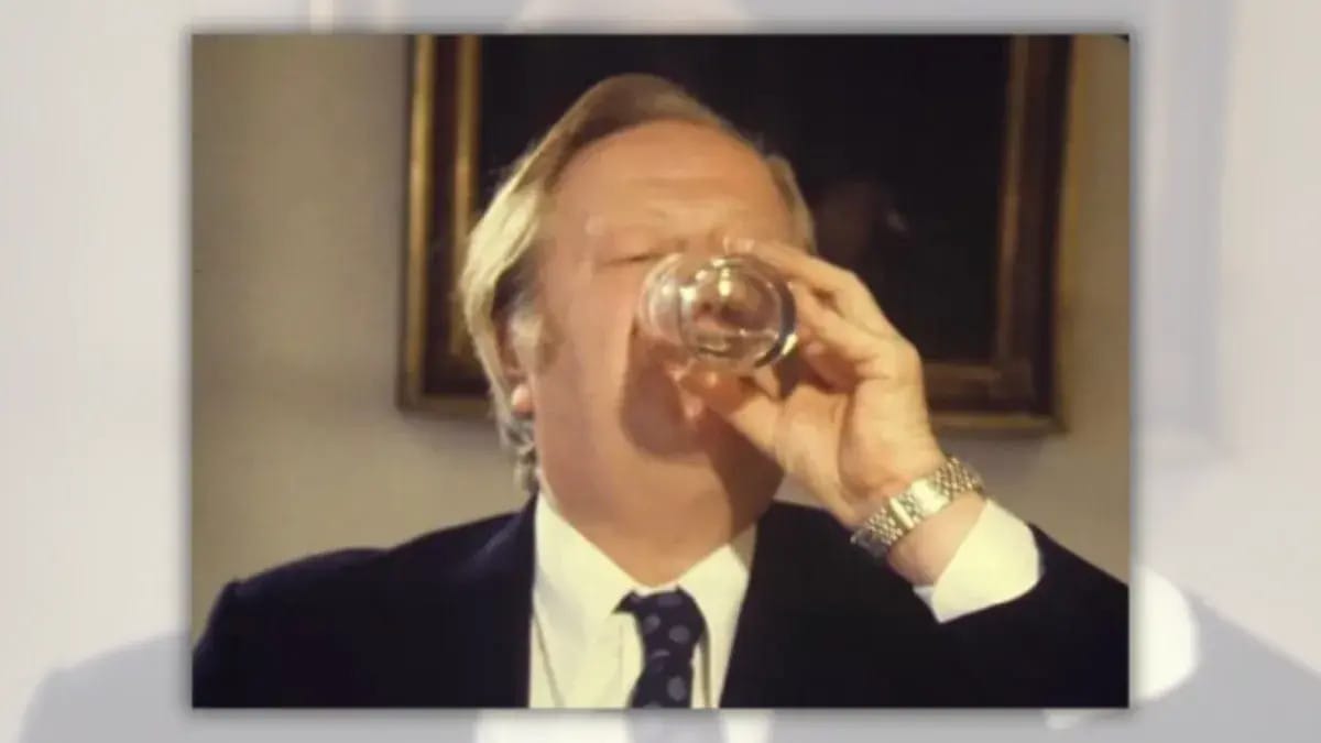 A white man wearing a black suit appears to drink some sort of liquid