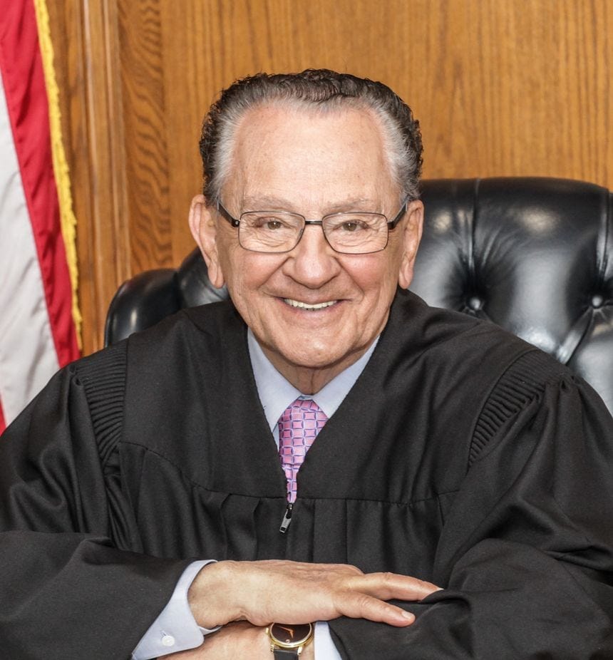 An older man in judicial robes sits smiling at a desk, with hands folded. He wears glasses, a light purple shirt, and a patterned pink tie. An American flag and wood paneling are visible in the background.