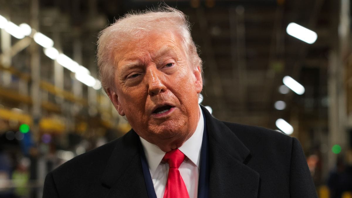 A white man wearing a black suit appears to speak while standing indoors.