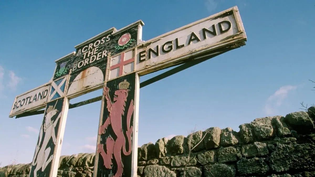 A gate says CROSS THE BORDER in front of a stone fence. On one side, the sign says SCOTLAND with a unicorn underneath, while the other side says ENGLAND with a lion underneath. The country's two flags are next to the respective words.