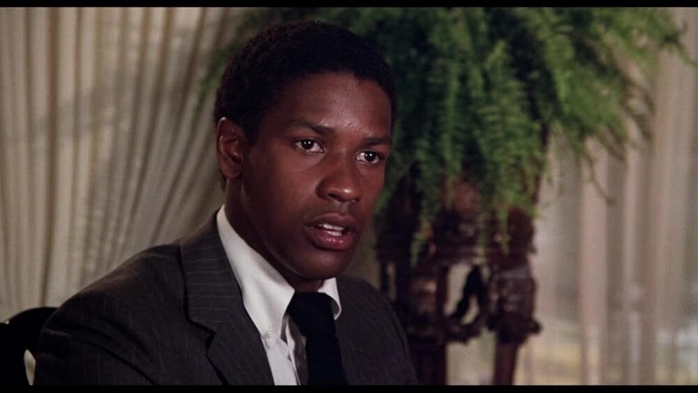 Screenshot of a young Denzel Washington in Carbon Copy, smiling in a button-down shirt, seated in a suburban home.