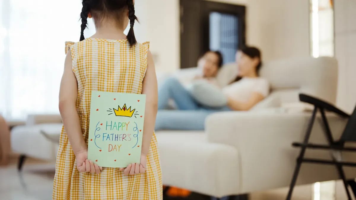 A kid holds what looks like a card that says HAPPY FATHERS DAY behind their back. They look towards two adults who are sitting on a couch, presumably their parents.