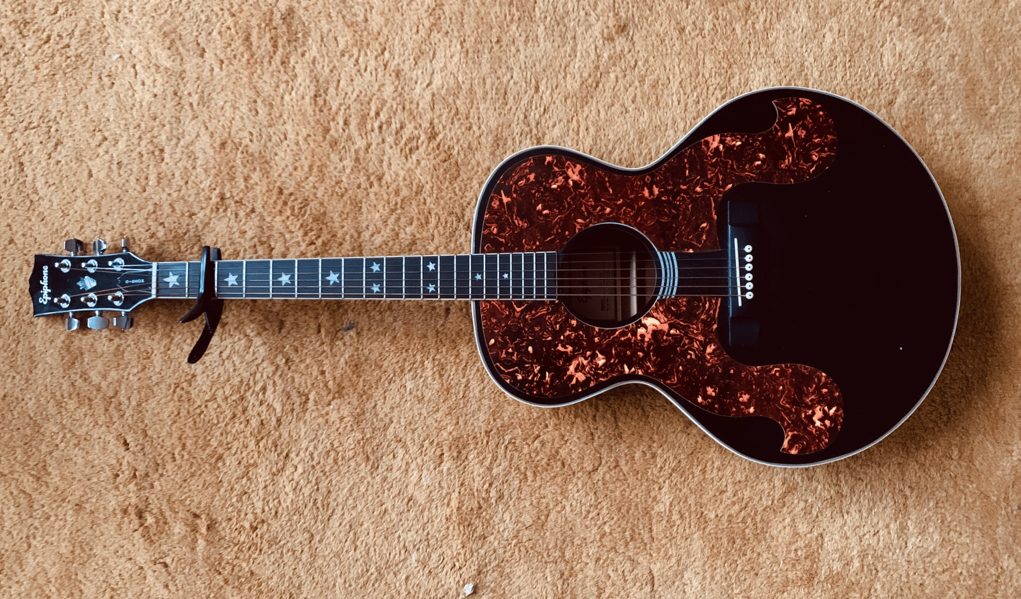 A photo of a six-string Epiphone knockoff of an Everly Brothers guitar sitting on a gold-colored carpet. The guitar has a black neck with inlaid stars, and the black body has an enormous and frankly overdone tortoiseshell pick guard. There's a capo on the second fret.