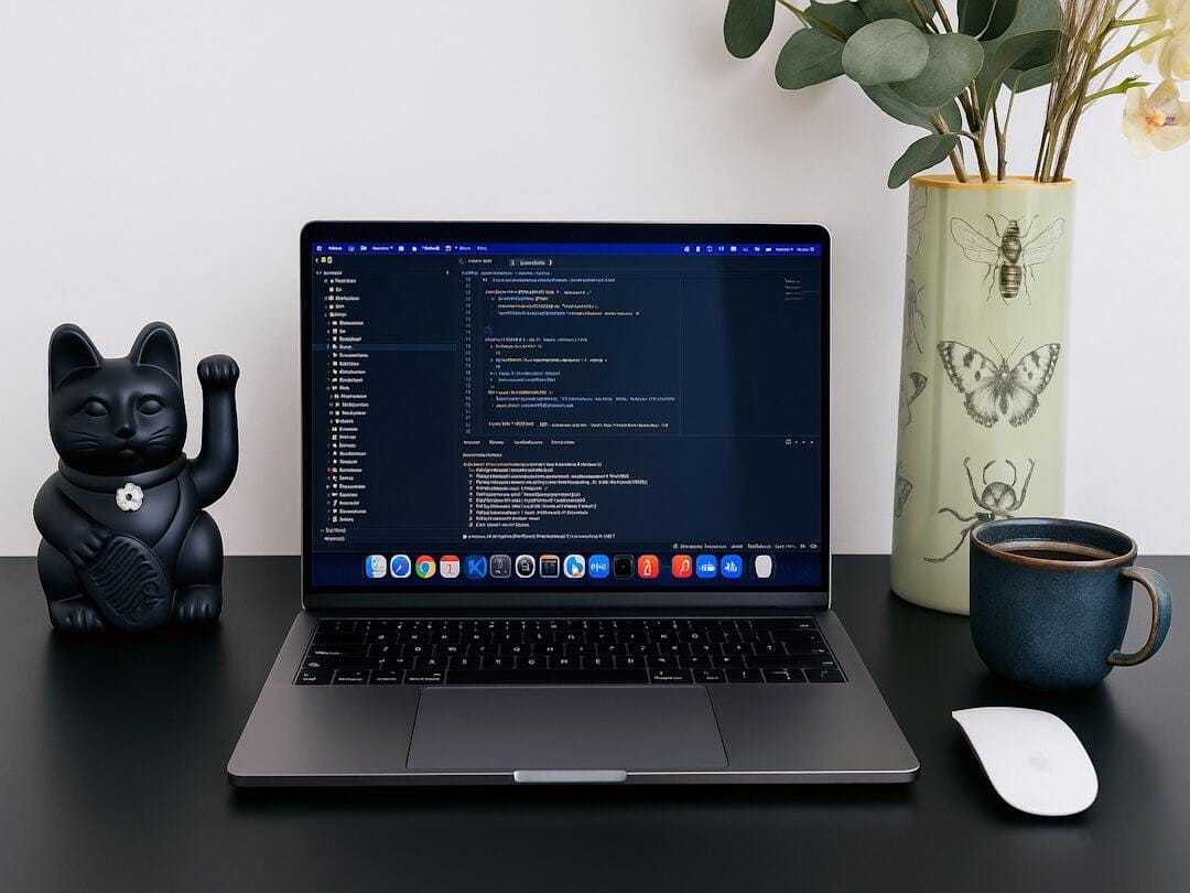 A sleek Apple MacBook Pro 2025 on a modern black desk setup, surrounded by stylish details including a matte black fortune cat, a botanical vase with plants and flowers, and a ceramic coffee cup. The screen shows coding in progress, highlighting themes of web development, design, and creativity. Perfect workspace inspiration for developers, designers, and digital creatives who value both productivity and aesthetics.
