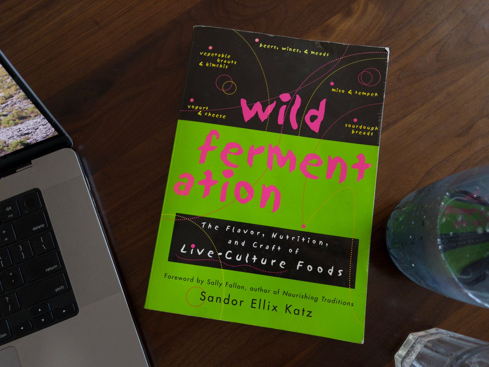 A photo of the book Wild Fermentation laying on a wooden table next to a computer and a glass of water.