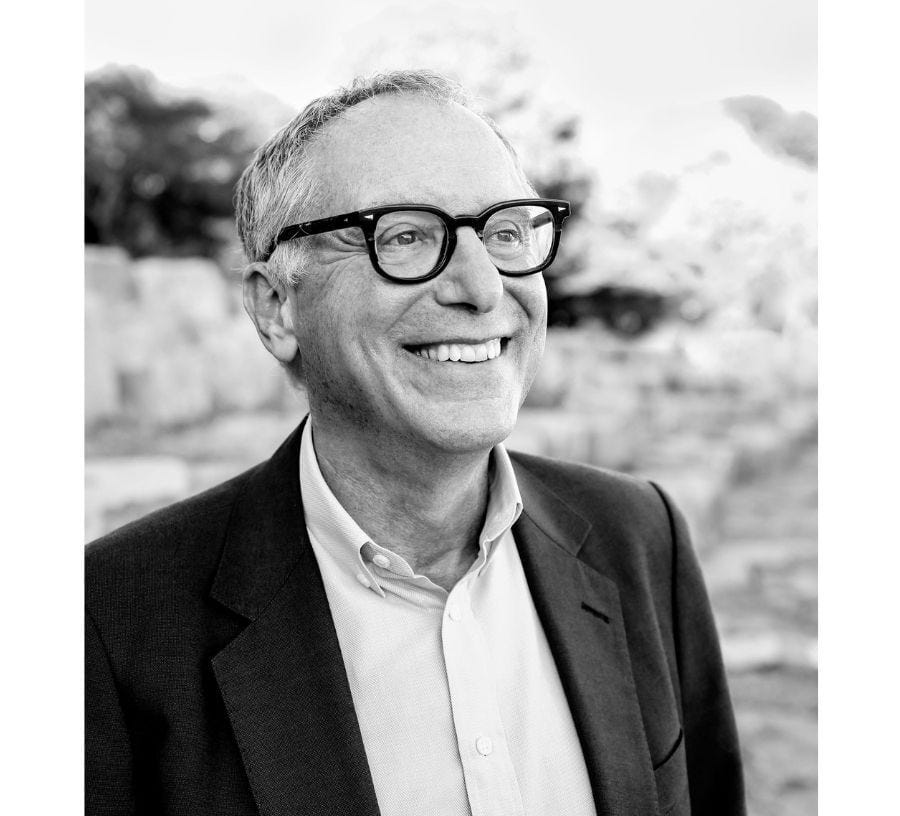 A grayscale portrait of a smiling man with short gray hair wearing dark-rimmed glasses, a suit jacket, and a collared shirt, looking slightly to the side with a blurred outdoor background.