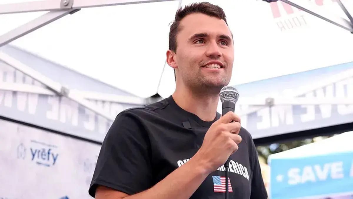 A white man smiles while holding a microphone. He appears to be standing under a tent.