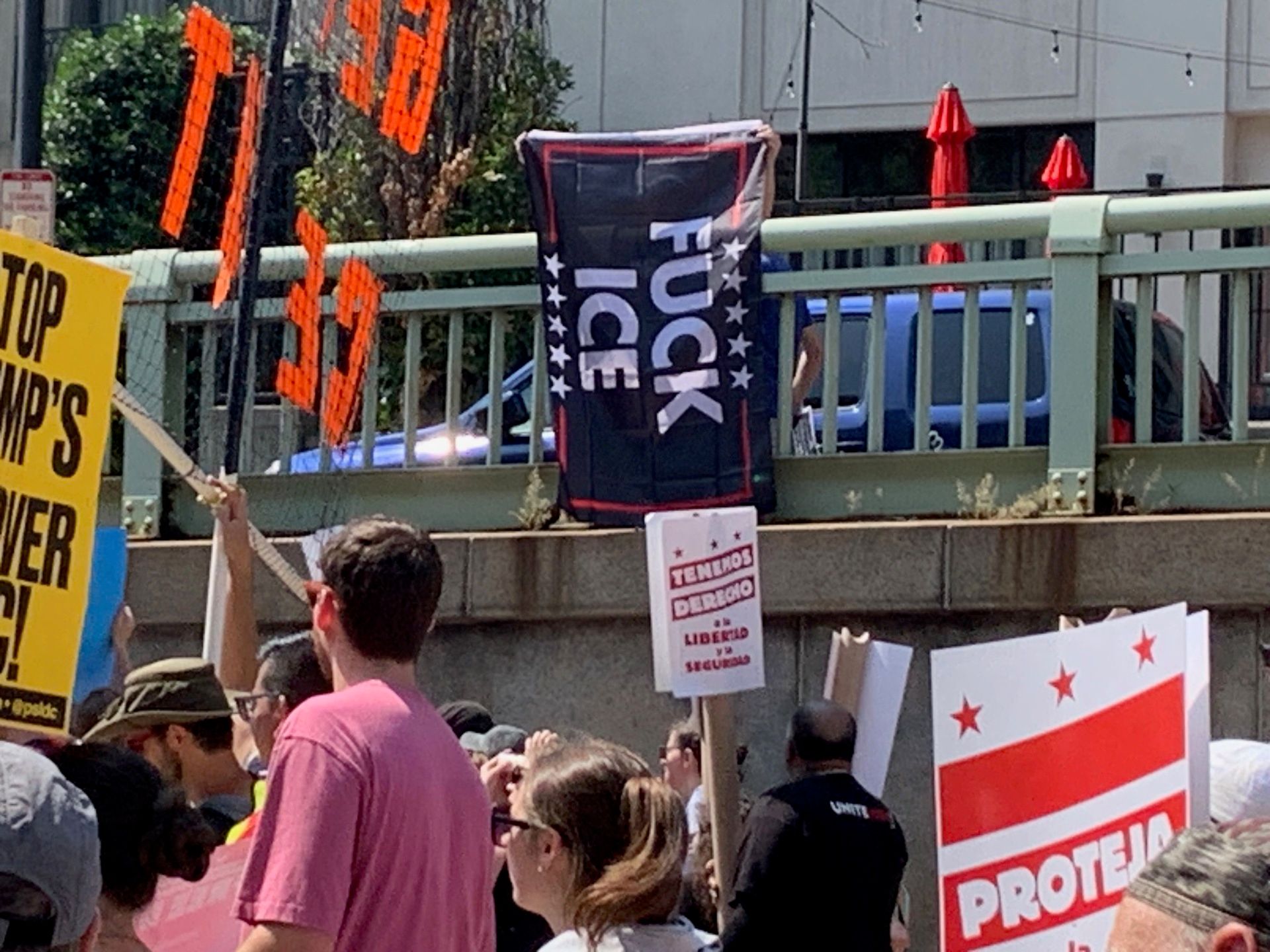 A crowd is visible, moving along the bottom of the photo, heading left. Above them, susspended from a railing, is a banner that reads "FUCK ICE."