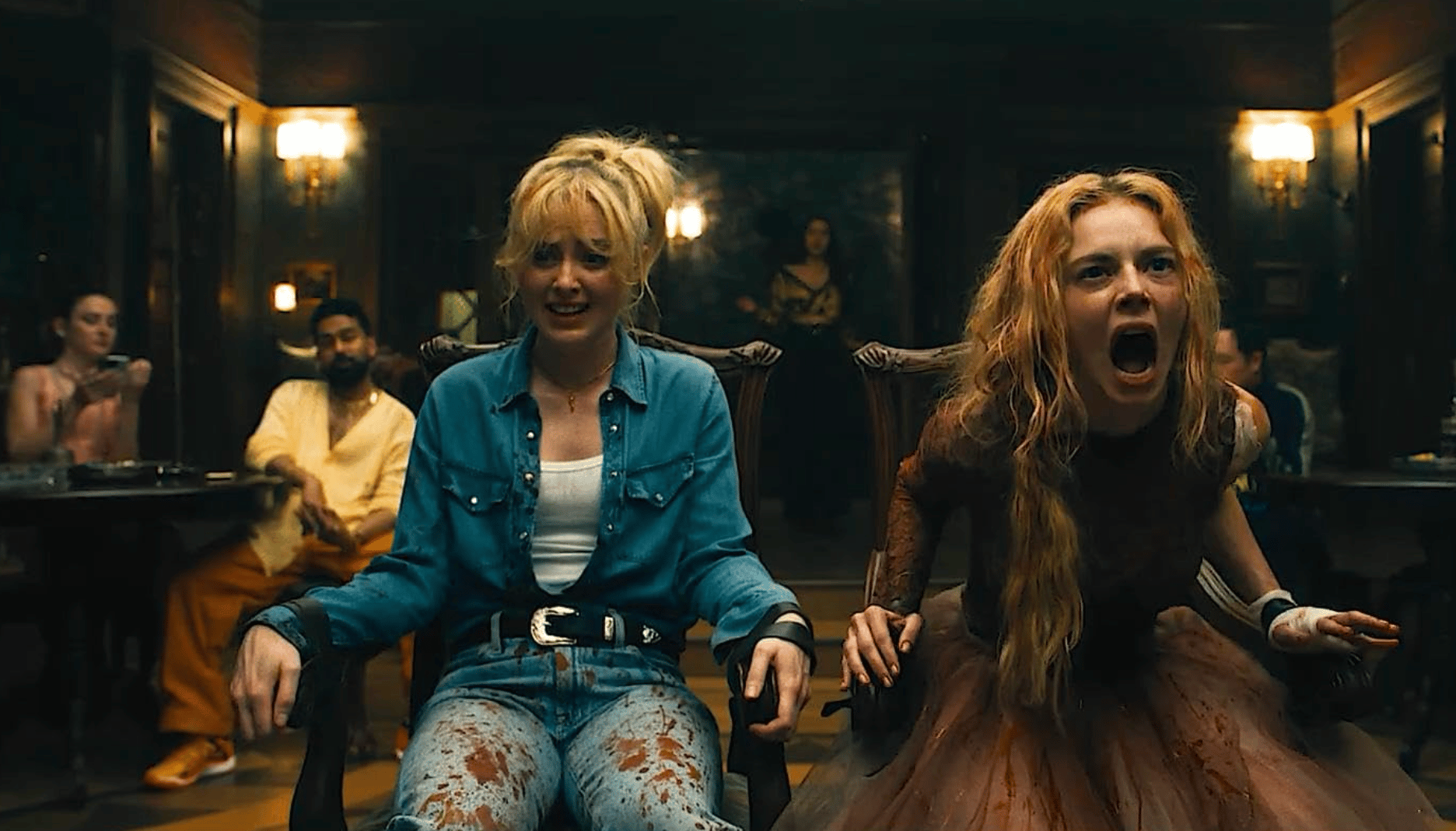 Two women are tied up in a pair of chairs in an ornate room. One is in a jean shirt and patterned leggings and looks about to cry. The other is in a bloody wedding dress and is screaming at something offscreen. Behind them a group of people sit, casually watching. 