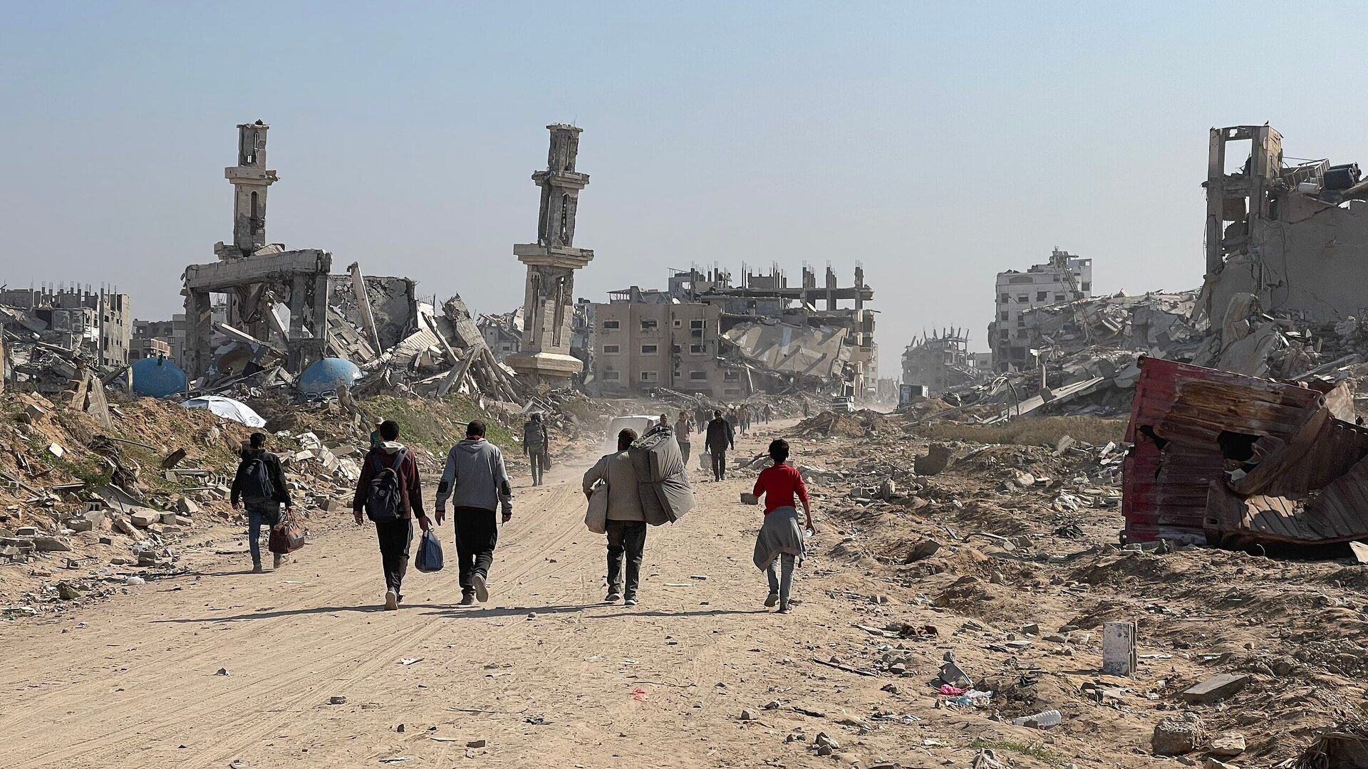 This photograph shows 5 people walking away from the camera on a dirt road during a sunny, cloudless day. They are in the mid-ground of the photo. Piles of rubble sit to the left and right of the road. In the background, there are more people walking down the same dirt road. To their left and right are the shells of destroyed buildings. DESCRIPTION FROM SOURCE: "Forced Displacement of Gaza Strip Residents During the Gaza-Israel War 23-25." CREDIT FROM SOURCE: Jaber Jehad Badwan. SOURCE: https://commons.wikimedia.org/wiki/File:Forced_Displacement_of_Gaza_Strip_Residents_During_the_Gaza-Israel_War_23-25.jpg. This file is licensed under the Creative Commons Attribution-Share Alike 4.0 International license.