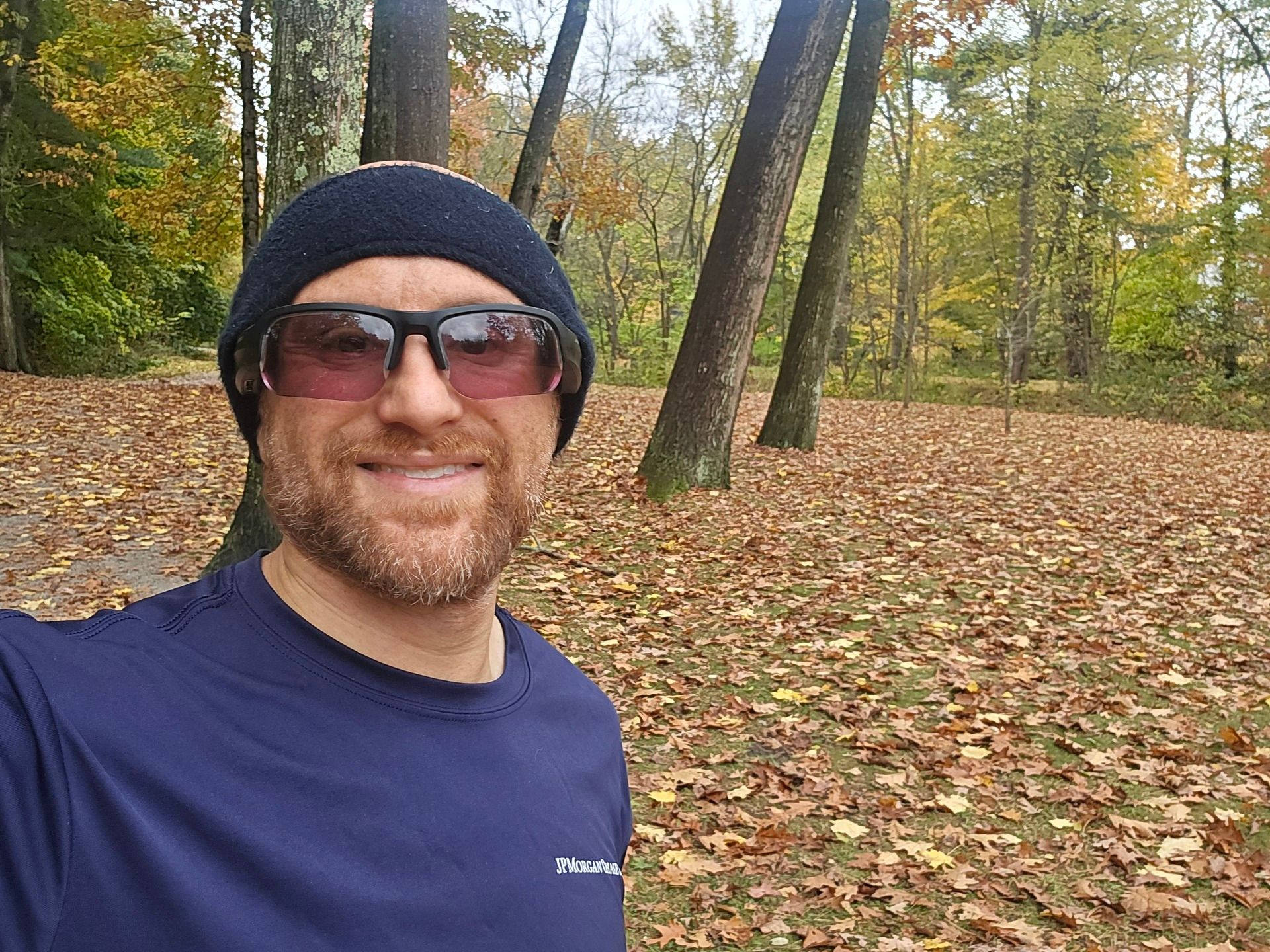 A selfie of me in front of a small field covered with fallen leaves, with forest beyond, on a bright day. I'm a bald, middle-aged, white man with a red beard flecked with white. I'm wearing a black headband, black sunglasses, and a long-sleeved dark blue running shirt with JPMorganChase written in white on the left chest.
