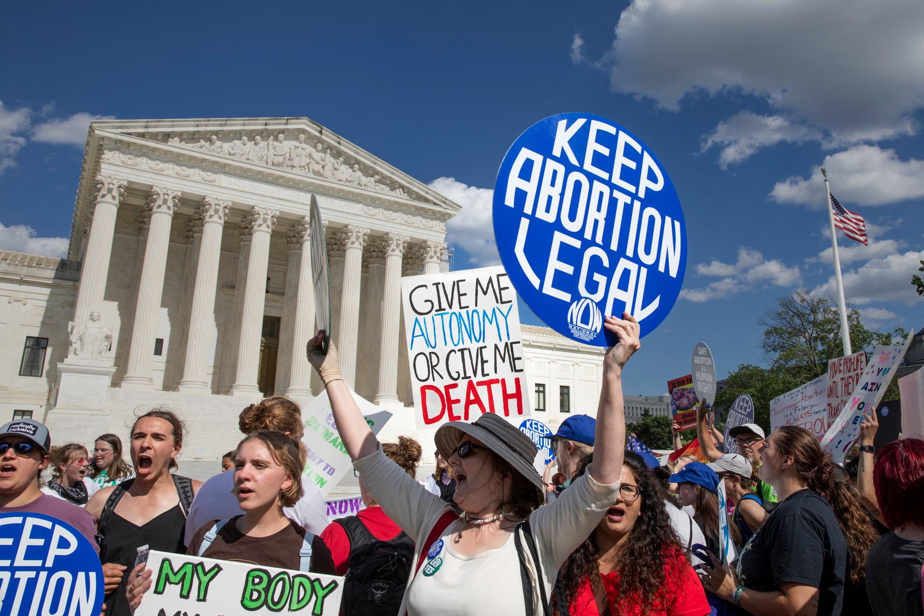 A group of abortion rights supporters protest outside the US Supreme Court. People hold signs reading “KEEP ABORTION LEGAL,” “MY BODY,” and “Give me autonomy or give me death.” The court building with its white columns is visible in the background under a blue sky.