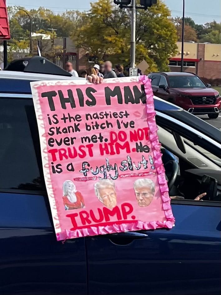 Someone in the passenger seat of a car holds an elaborately decorated glittery sign with the text, "This man is the nastiest skank bitch I've ever met. Do not trust him. He is a fugly slut! Trump."