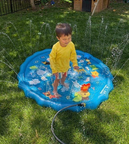 Young boy laughing and running through a splash pad with water spraying around him on a sunny day.