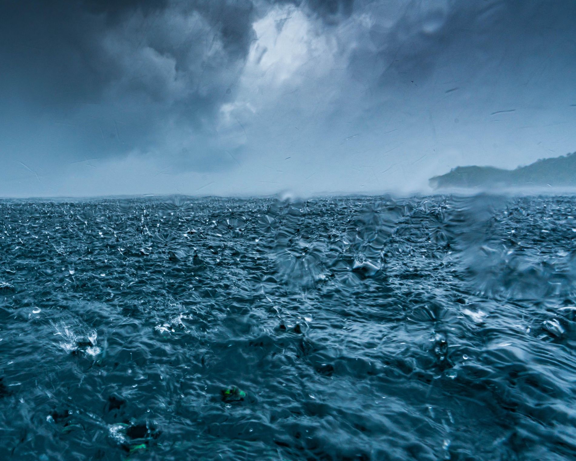 Dark storm clouds loom over a restless ocean as heavy rain pelts the water’s surface, creating ripples and splashes; a distant shoreline is faintly visible through the mist.