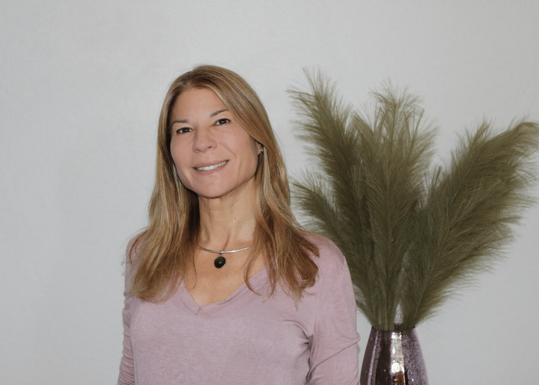 Lisa smiles in front of a vase of palm fronds. She has shoulder length hair and wears a lilac shirt.