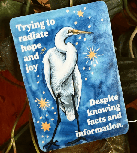A drawing of a white egret on a blue background with yellow stars. White lettering reads: “Trying to radiate hope and joy despite knowing facts and information.