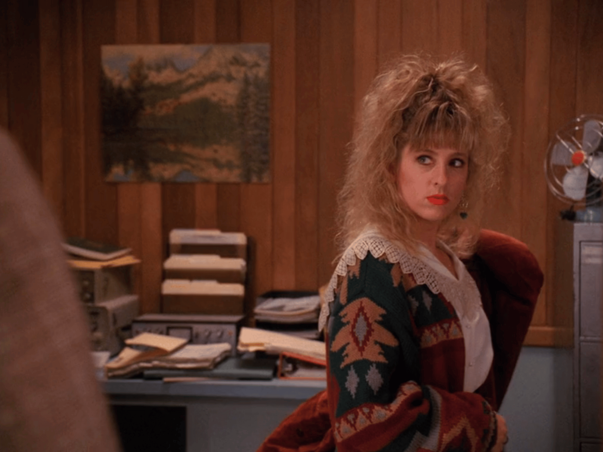 Kimmy Robertson as Lucy Moran in TWIN PEAKS. She is wearing an Aztec-style cardigan that is green, red, gold, and gray. She also has a white blouse with a ruffled collar.