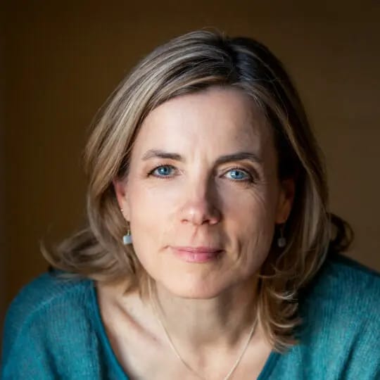 A woman with light brown hair and blue eyes looks directly at the camera with a calm expression. She is wearing a teal sweater, small earrings, and a delicate necklace, with a soft brown background behind her.