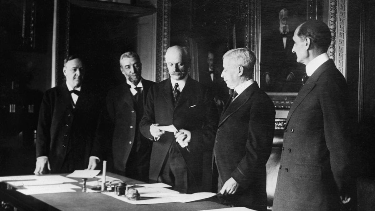 A black and white photo shows five people standing around a table. The person in the center is holding a slip of paper in their hands, being looked at by the four members gathered around them. They stand behind a table that has paperwork on it.