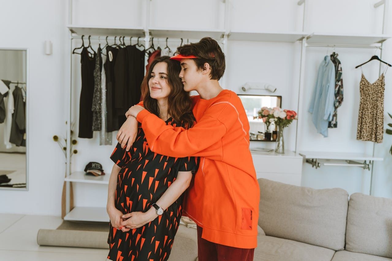 Teen hugging their mother from behind in a cozy, stylish home setting filled with hanging clothes and flowers.