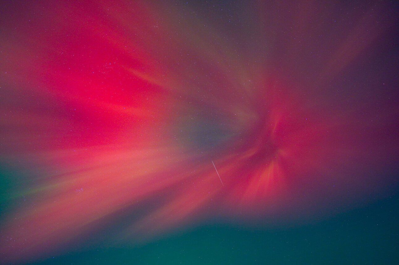 A photo of the aurora borealis. A dark sky peppered with tiny white stars. Over the top is a huge, vivid swirl of vibrant pink as the northern lights radiate outwards. Just about visible is a tiny streak of light in the middle: a shooting star against this stunning scene. 