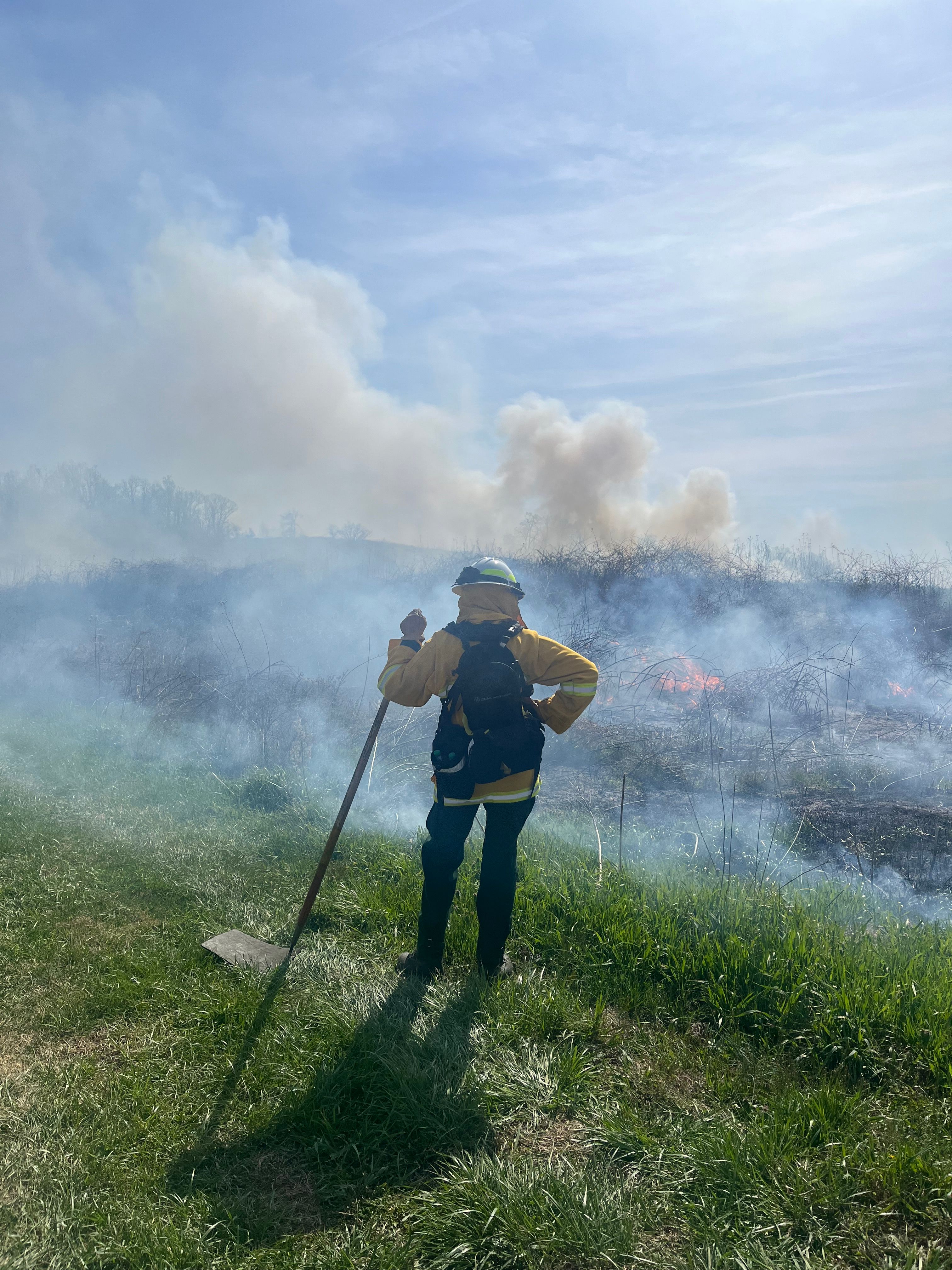 a volunteer in PPE looks out at the horizon while watching and monitoring the fire. Smoke billows up into a pale blue sky