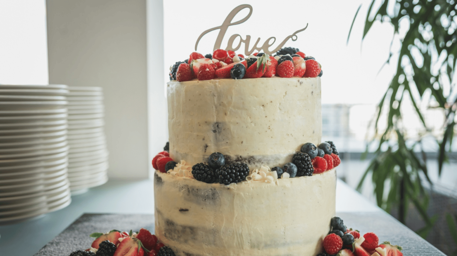 Close up of a wedding cake on a counter. The cake has minimal icing and the top is covered in various berries. There are more berries on the other layers. The cake sign reads, "Love"