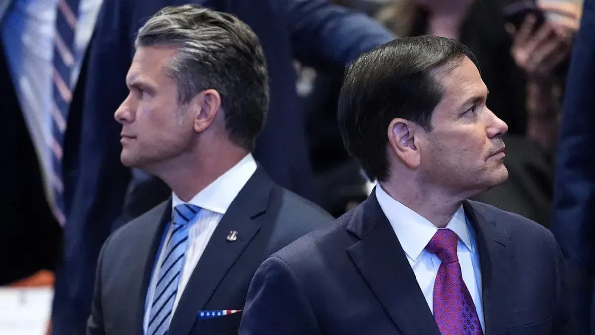 A white man wearing a dark blue suit looks towards the left. To his right, a Latino wearing a dark suit looks towards the right. They appear to be standing in front of a crowd of people.