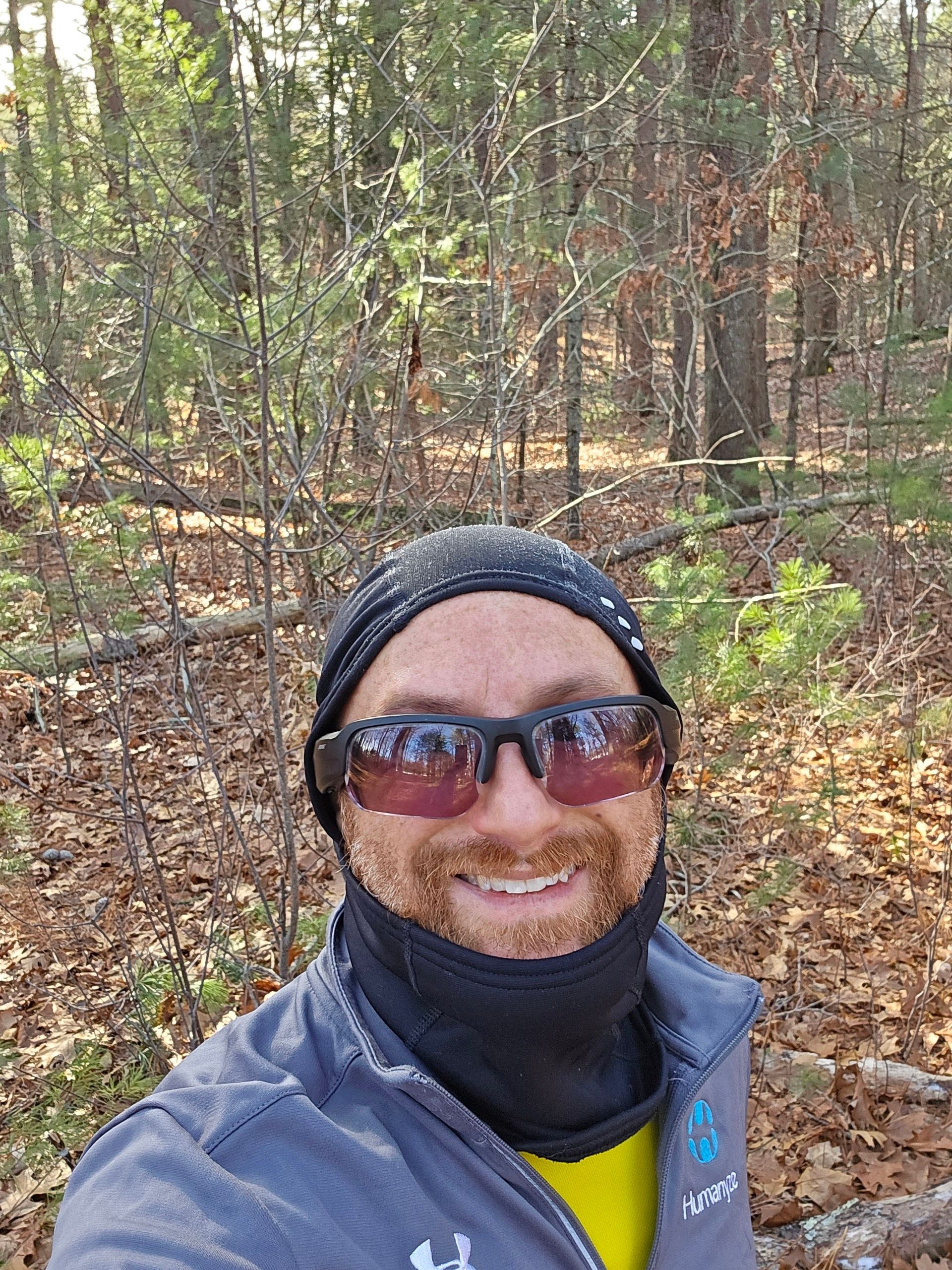 A selfie of me in a forest on a sunny day. I’m a middle-aged white man with a red beard flecked with white. I’m wearing a black balaclava with the mouth cover pulled down, black sunglasses, and a grey Under Armour partially unzipped sweatshirt with the Humanyze logo on the right.