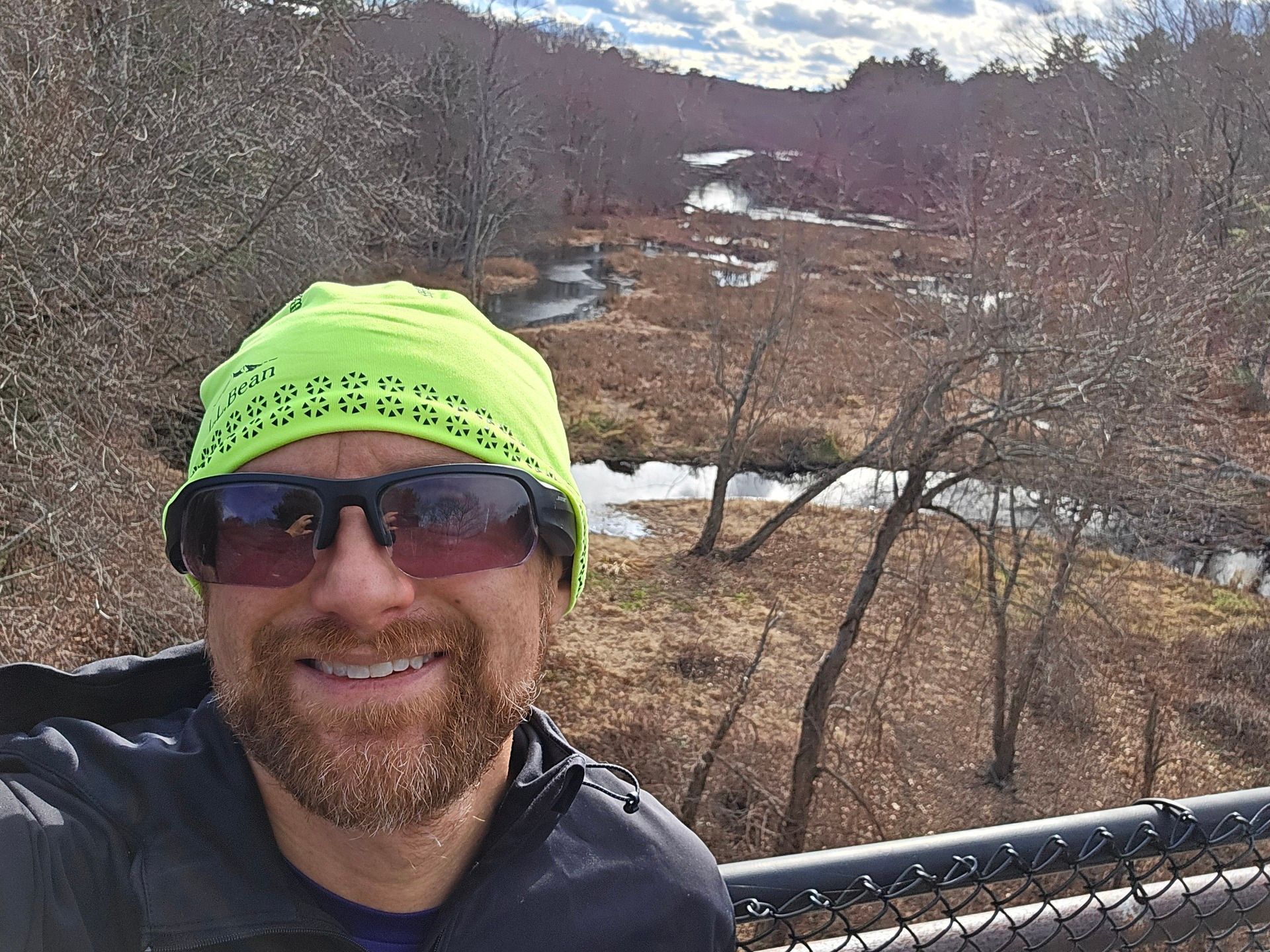 A selfie of me on a bridge above a mostly dry swamp, with a winding brook feeding into the larger Charles River beyond, on a bright, slightly cloudy day. Thick forest lines the banks of the swamp and river. I'm a bald, middle-aged, white man with a red beard flecked with white. I'm wearing a neon green LL Bean beanie, black sunglasses, and a black running jacket.