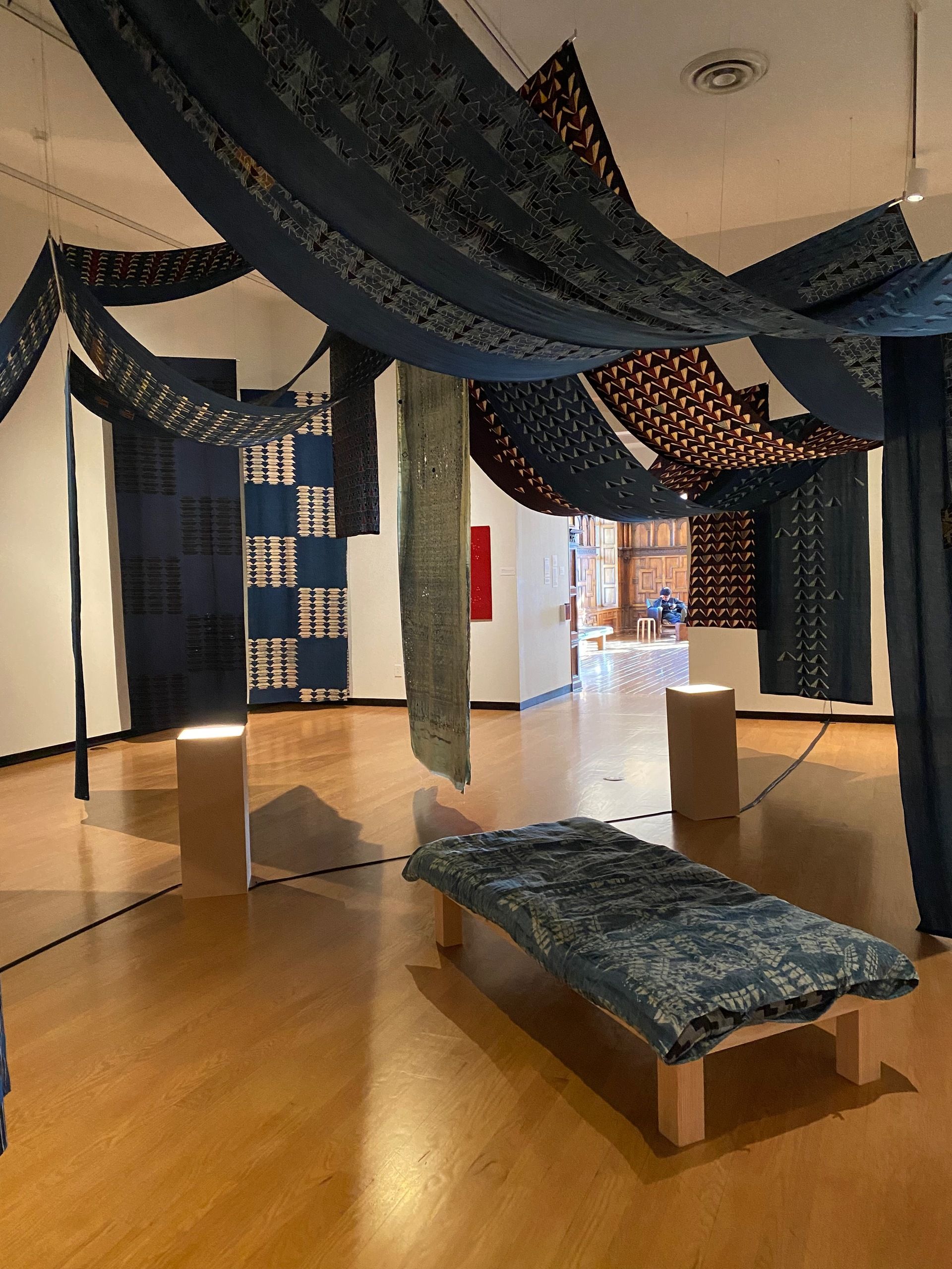 View of a large installation of block-printed textiles in shades of blue, red, white, and gray, in various patterns, hanging down from and across the ceiling. There is a long cushioned bench in the middle of the installation, under the textiles. 