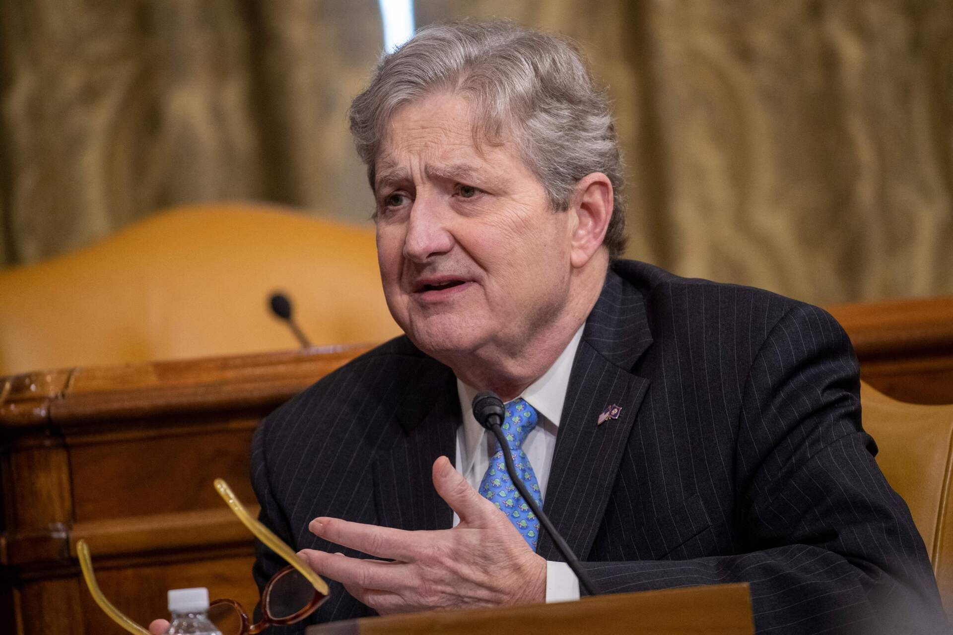 Close up of a white man wearing a black suit speaking at a podium. Part of his hand is visible at the bottom of the image