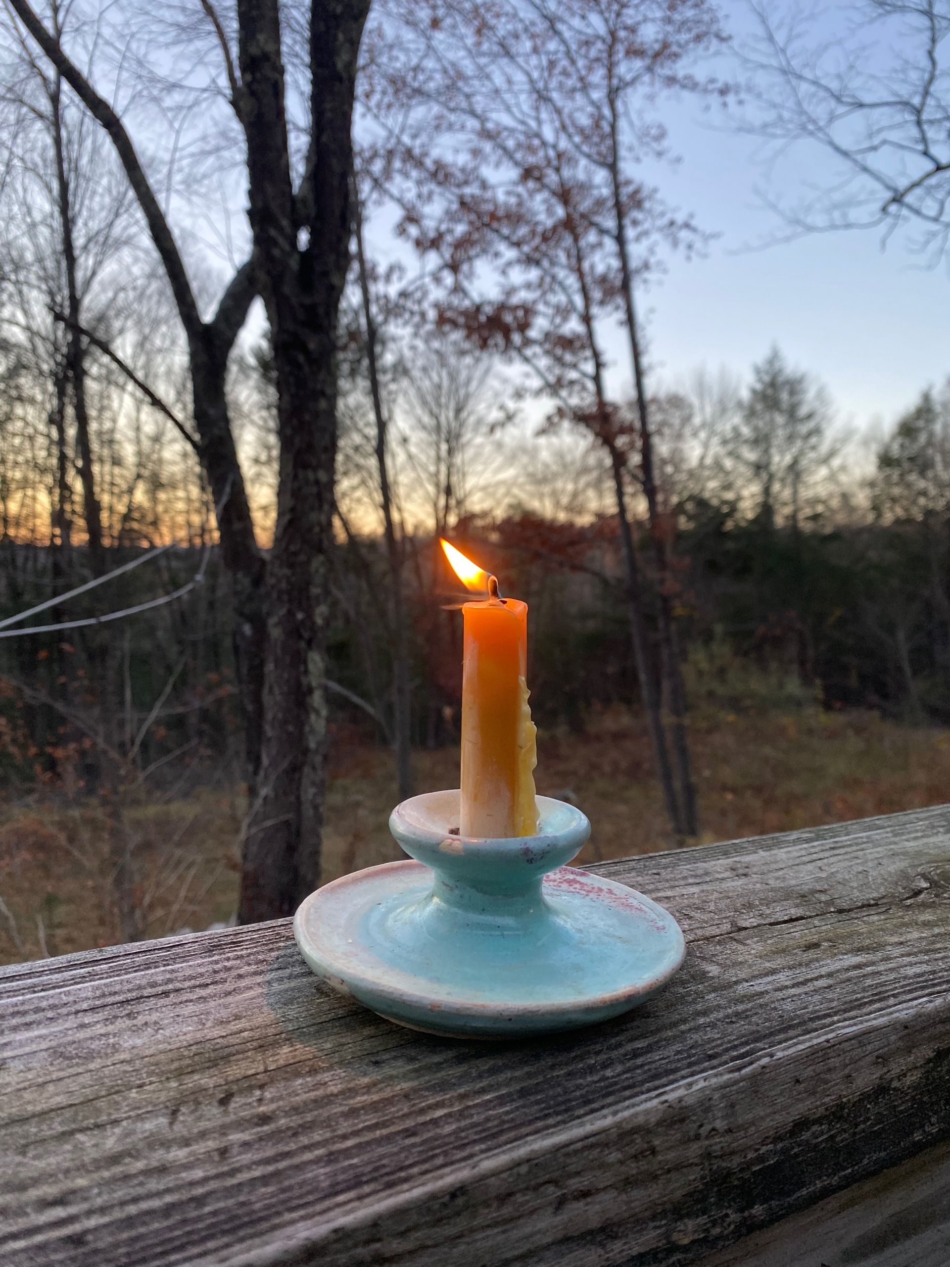 A lit candle in a ceramic candle holder sitting on a porch railing at twilight, in front of a blue and gold sky.