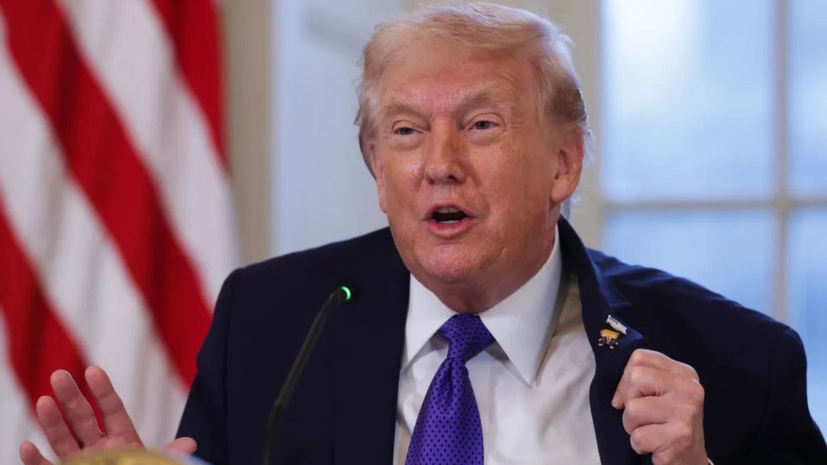 A white man wearing a dark blue suit speaks indoors. With his right hand, he holds the suit jacket as to draw attention to pins he's wearing there.