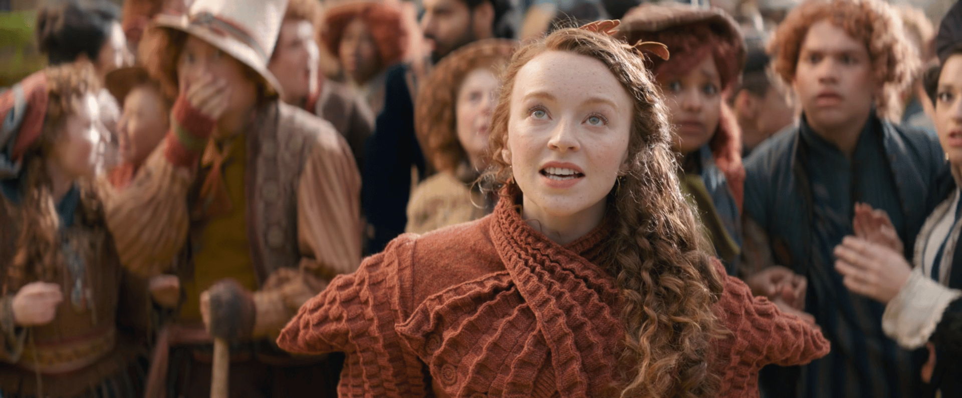 Kirsty Anne Shaw as Skeptical Munchkin in WICKED. She is wearing an orange frilly shawl sweater-type top. And yes, Skeptical Munchkin is literally her credit.