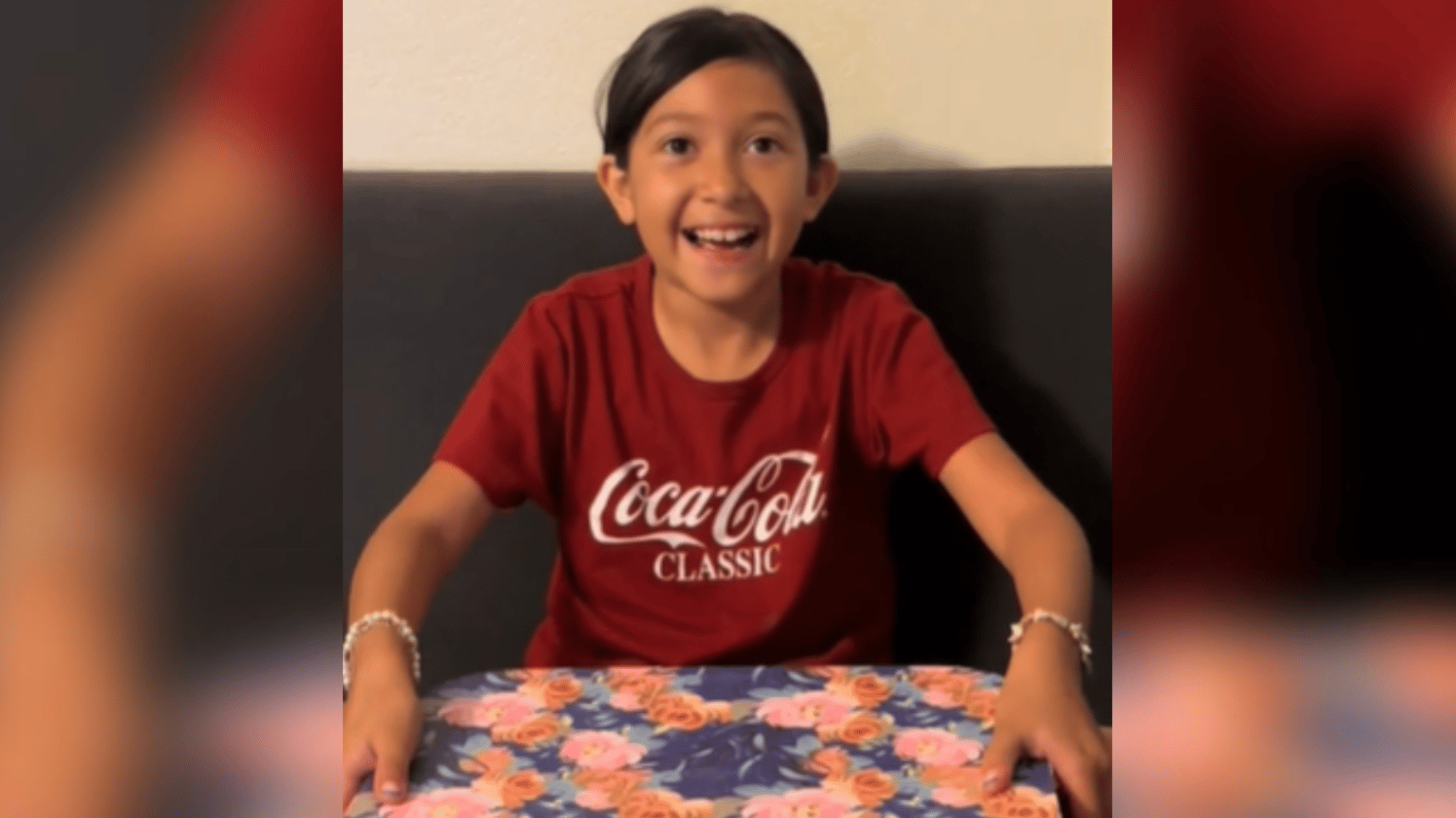 A little girl sits on a couch with a colorful box in front of her. Her eyes are wide in excitement and she's got a huge grin. She's starting to lift the lid off the box