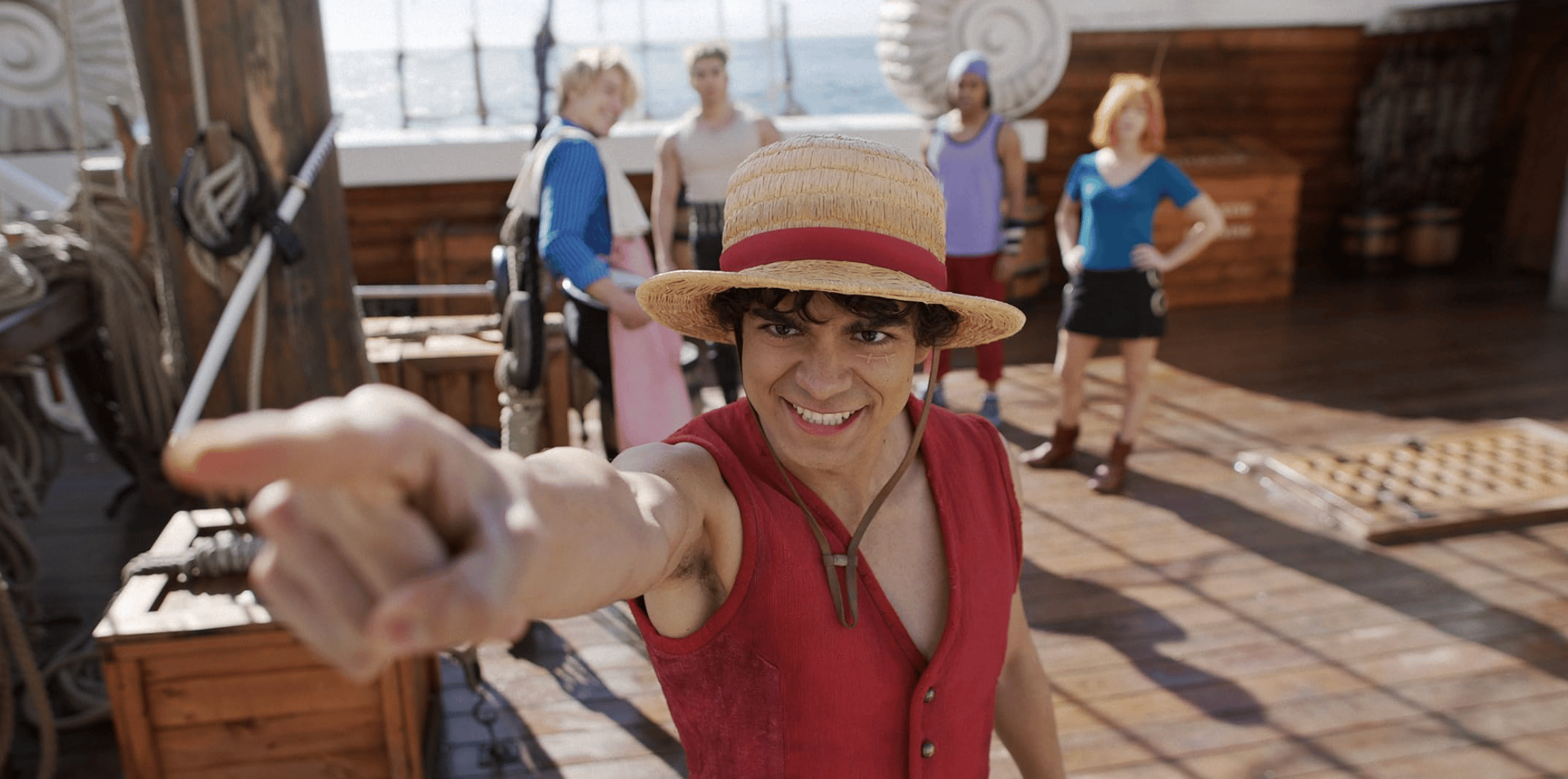 A young man in a red vest and a straw hat stands on a wooden ship deck and points into the distance, hinting at future adventures. Behind him a crew of fellow pirates (slightly burred) watch him while smiling.