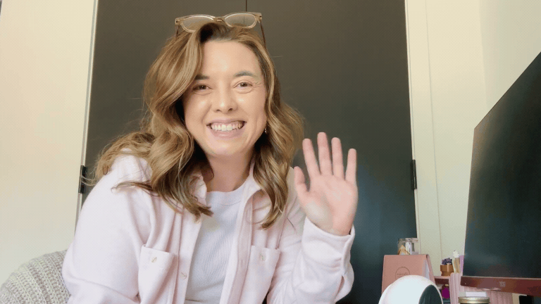 GIF of a woman in a light pink long sleeve shirt smiling and waving at the camera 