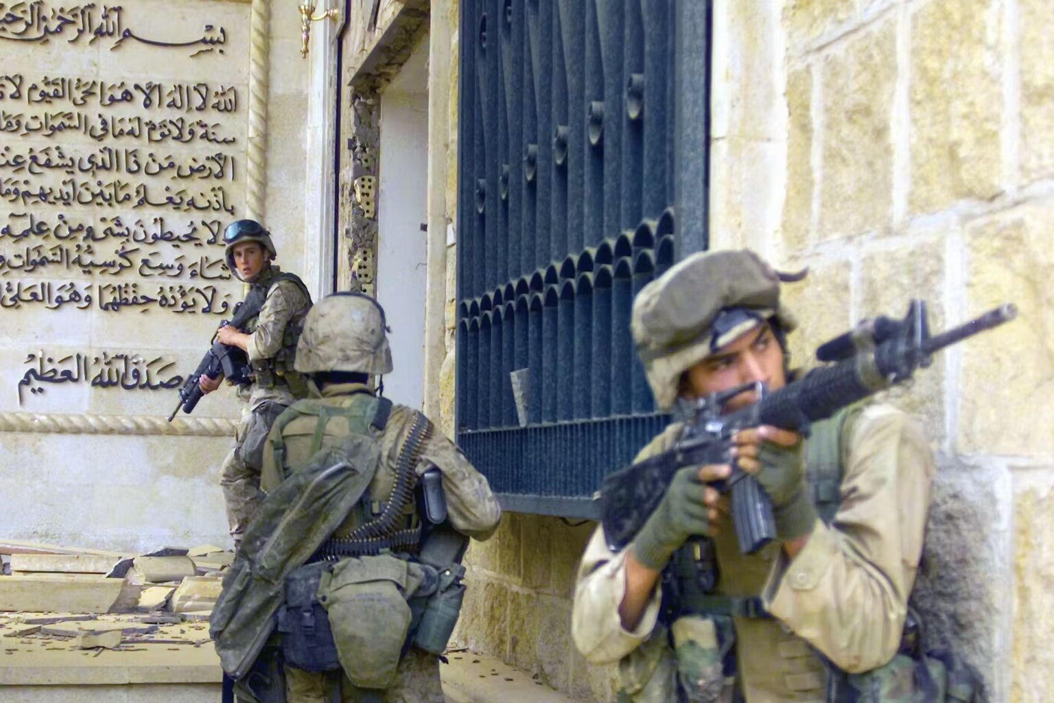 A photo of U.S. armed military personnel next to a high pale brick palace wall, with Arabic script on another wall behind them and wrought iron visible to one side.