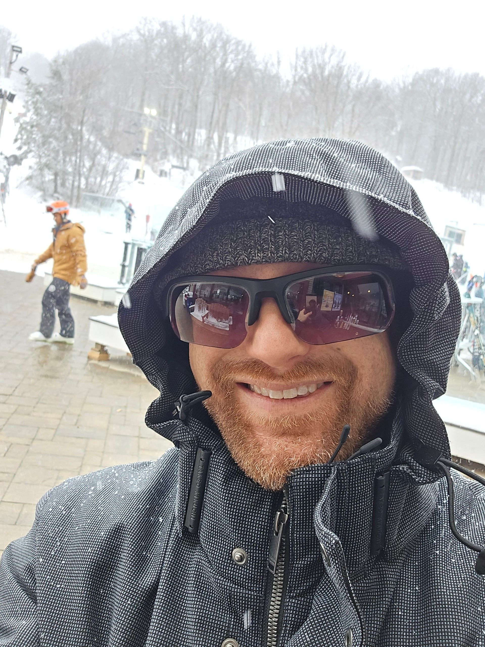 A selfie of me on a stone patio with people in ski gear behind me in front of a snow-covered mountain with a chair lift on the left. I'm a middle-aged white man with a red beard flecked with white. I'm wearing a blue winter coat with a dark blue graph check pattern with the hood pulled up, a grey winter hat, and black sunglasses.