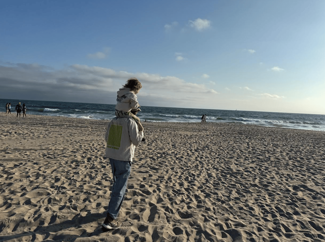 "An adult walks along a sandy beach near the ocean, carrying a young child on their shoulders. The sky is mostly clear with a few scattered clouds, and small groups of people are visible in the distance near the shoreline. The ocean waves gently meet the shore under late afternoon sunlight."